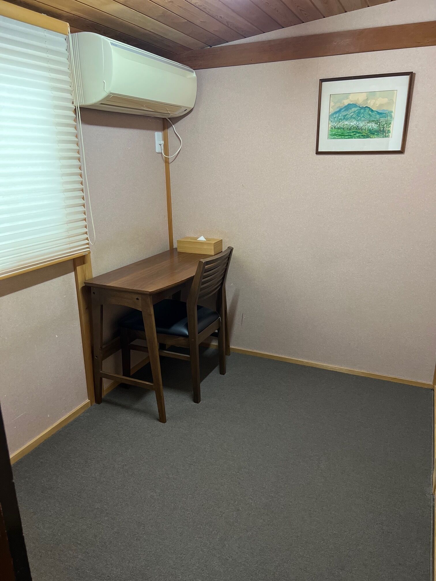 A corner room with a wooden table and chair, an air conditioning unit mounted on the wall, and a framed painting on the wall.