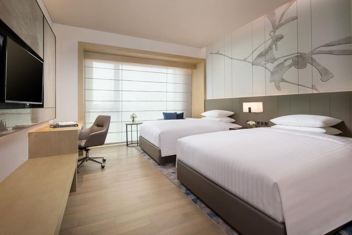 Ein Hotelzimmer mit zwei Betten, einem Schreibtisch, einem Sessel, einem Fernseher und einem großen Fenster.