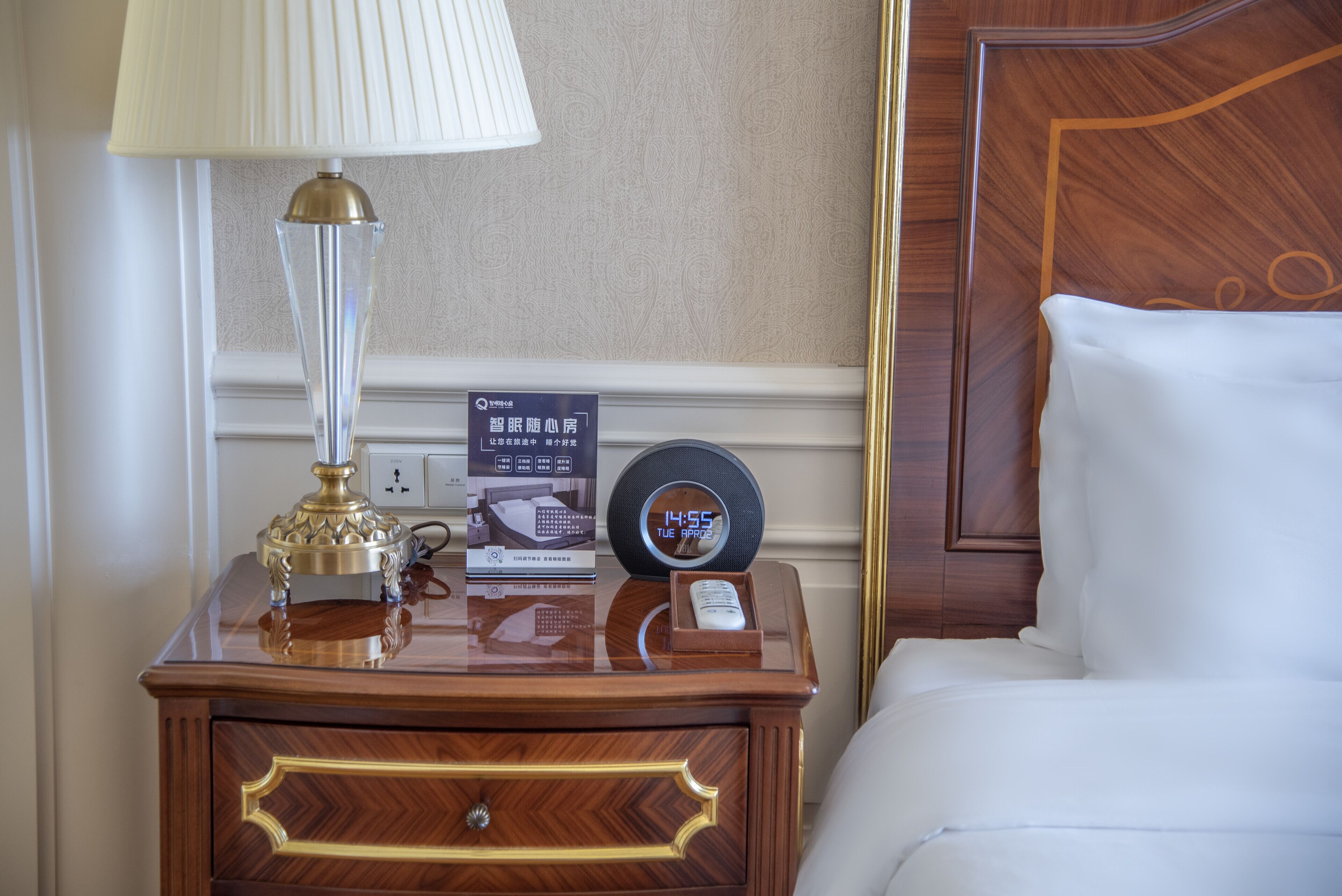 Une chambre d’hôtel bien rangée, avec une table de chevet en bois, une lampe, une horloge numérique et une télécommande.