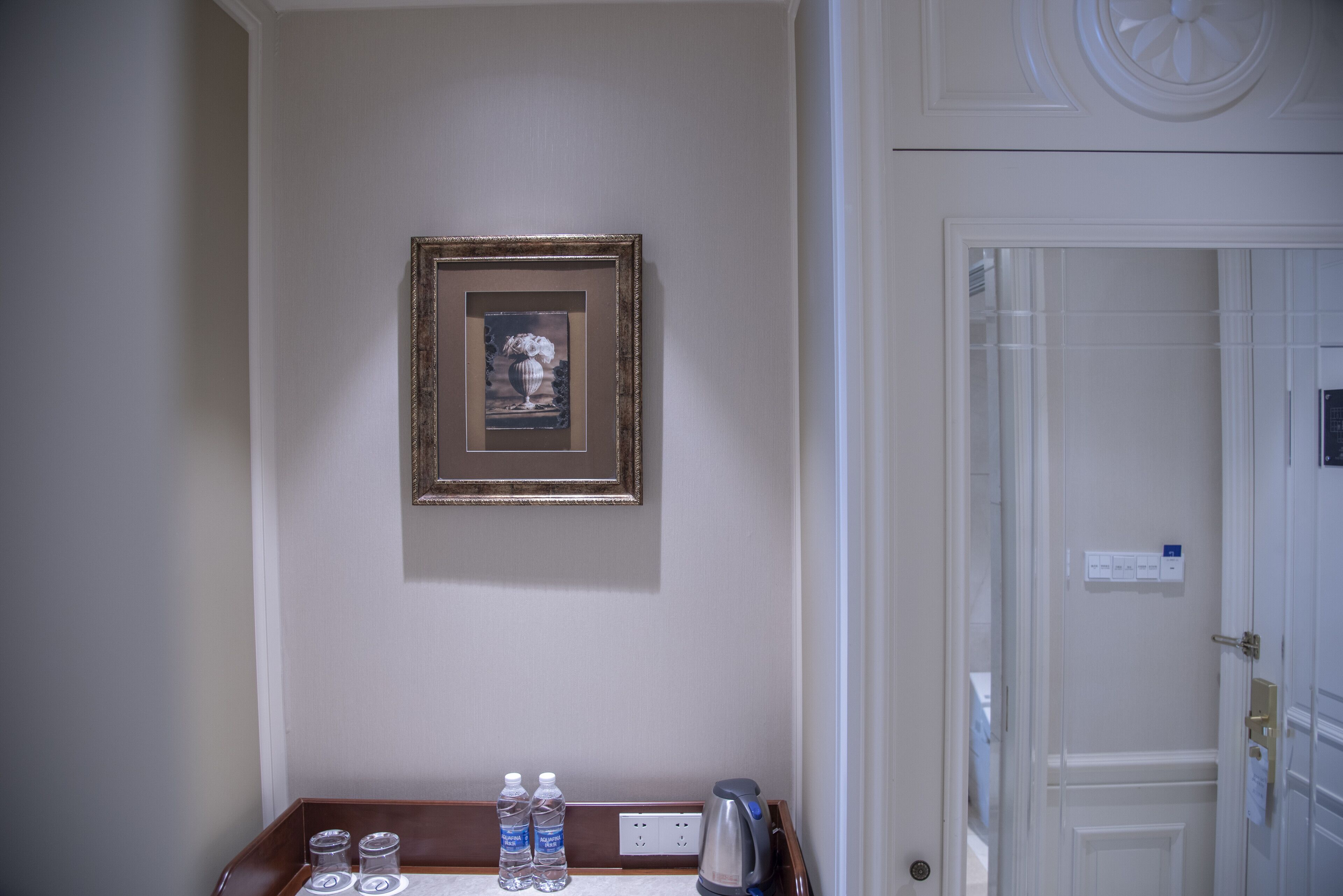 Ein Hotelzimmer mit einem gerahmten Bild an der Wand, einem kleinen Tisch mit Wasserflaschen und einem Wasserkocher und einer Tür mit einem Schild.