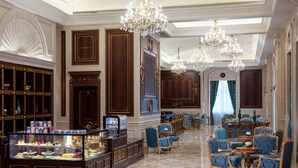 Un hall d’hôtel luxueux, agrémenté de boiseries sculptées, d’un lustre central et d’une vitrine où sont exposés des desserts.