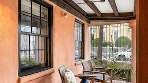 A cozy outdoor seating area with wicker chairs and a small table, overlooking a street with parked cars and a white fence. - Charming Historic 1BR w/ Private Porch + Free Parking (Charleston)