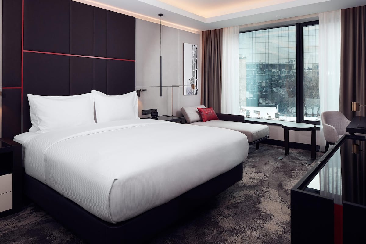Habitación de hotel moderna con una cama grande, un escritorio, una silla y vistas a la ciudad a través de amplias ventanas.