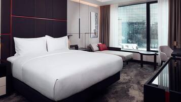 A modern hotel room with a large bed, a desk, a chair, and a view of the city through large windows.