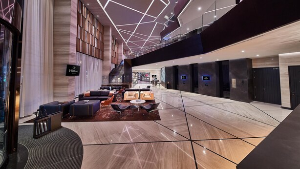 A spacious lobby with a modern design, featuring a seating area with sofas and armchairs, a staircase, and a 'Hilton' sign.