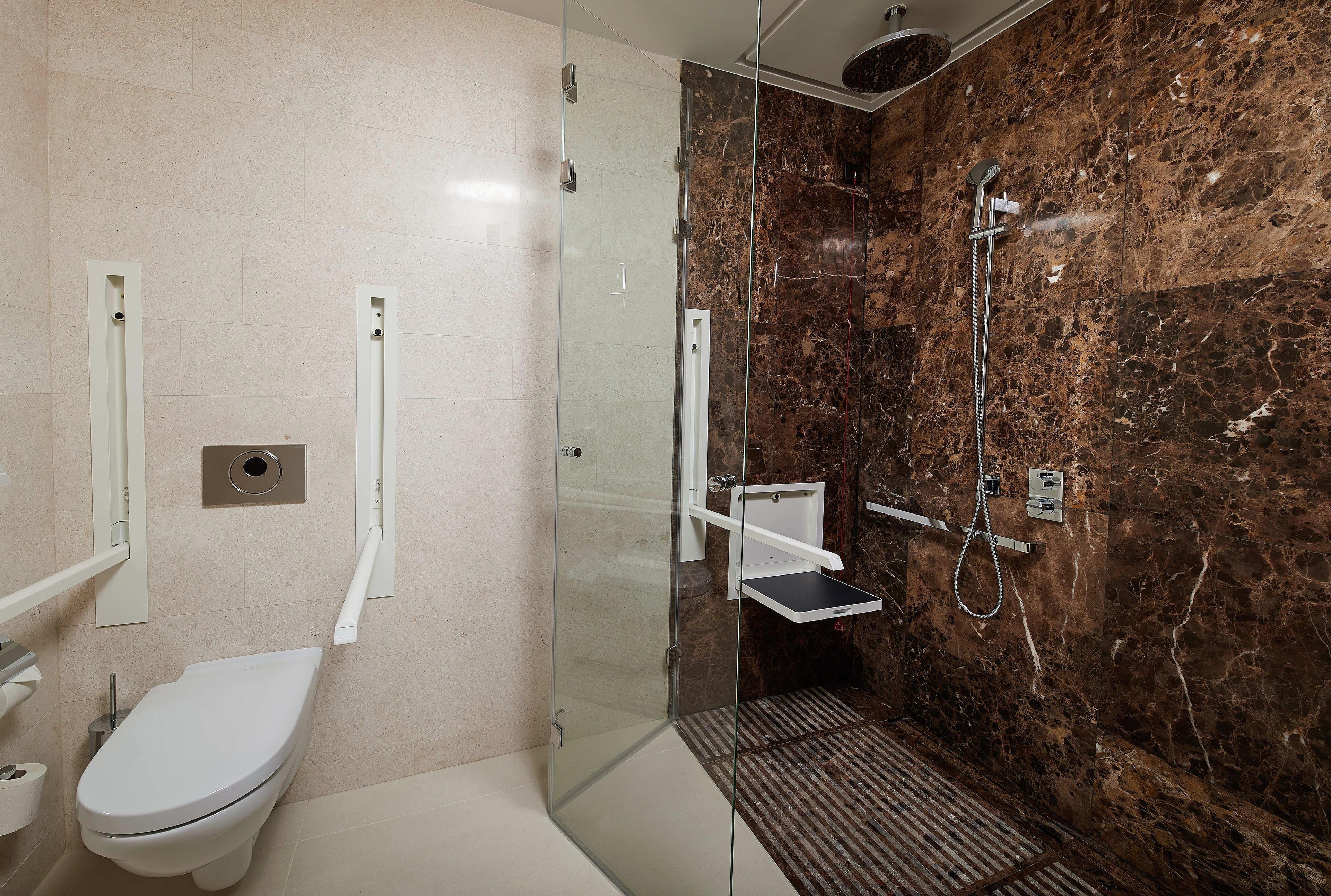 A bathroom with a toilet, a glass-enclosed shower with a marble wall, and a small shelf.