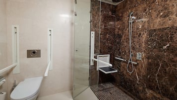 A bathroom with a toilet, a glass-enclosed shower with a marble wall, and a small shelf.