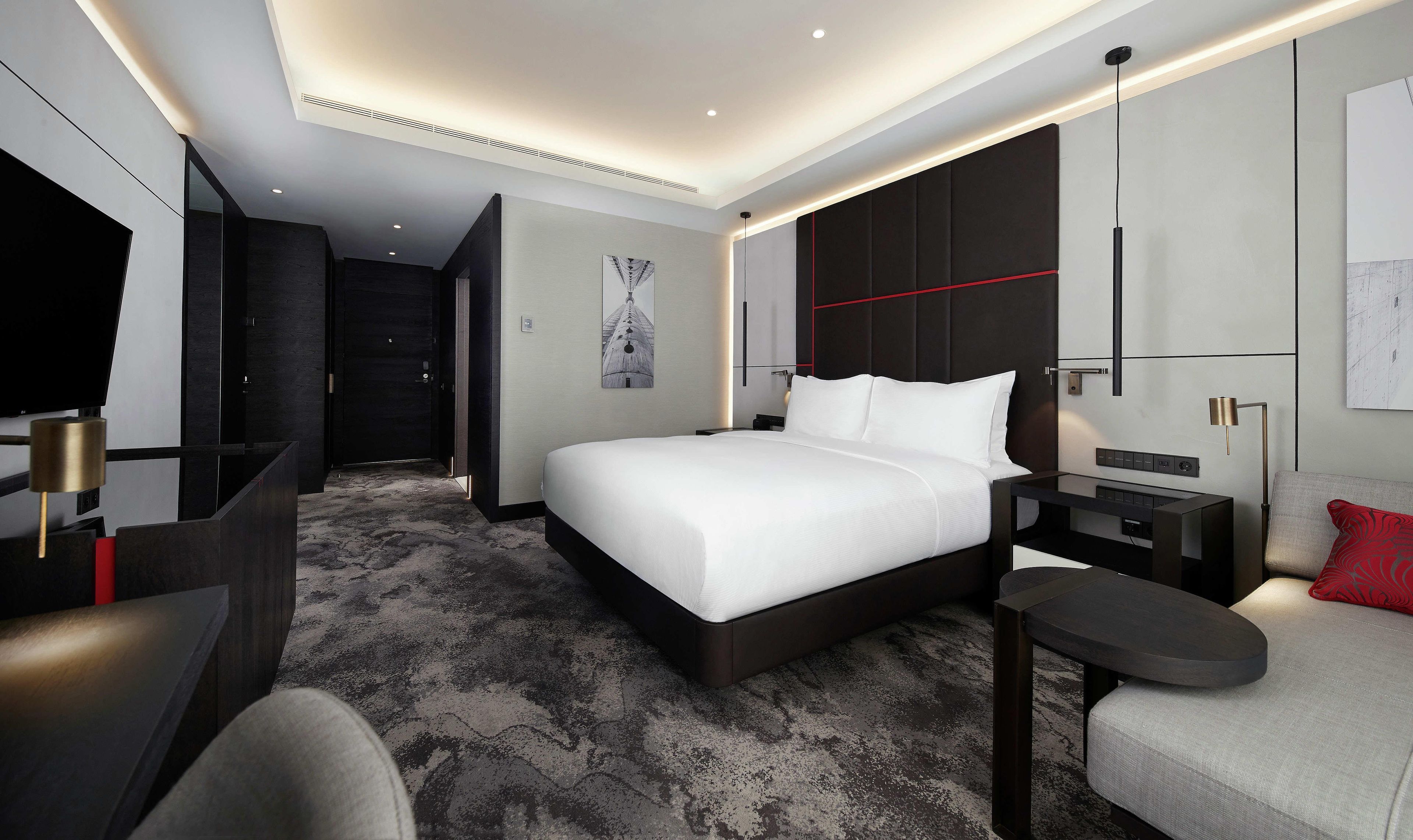 Ein modernes Hotelzimmer mit einem großen Bett, einem Schreibtisch, einem Stuhl, einem Fernseher und einem roten Zierkissen.
