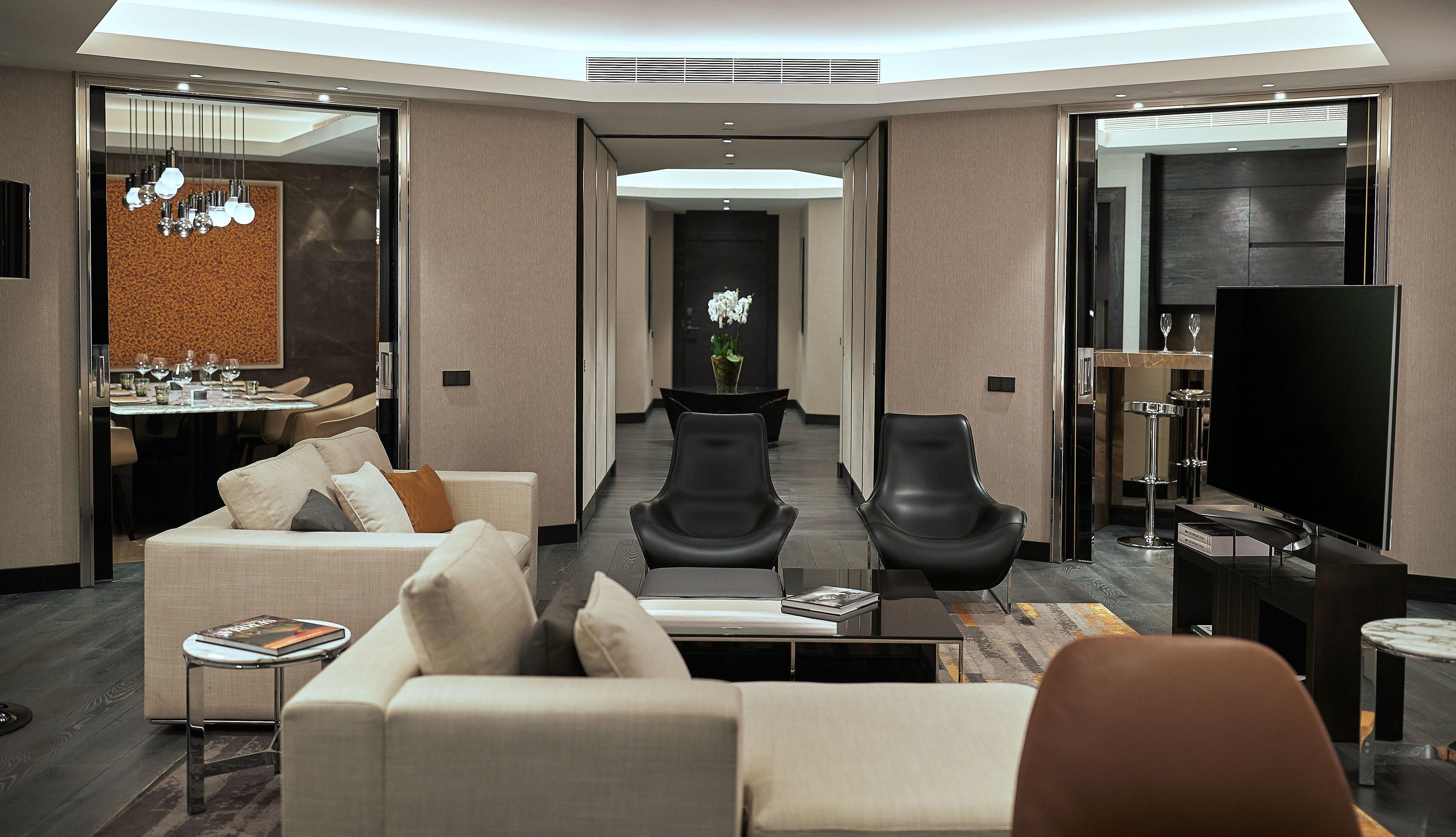 A modern hotel room with a sofa, two chairs, a TV, a dining area, and a hallway leading to a door.