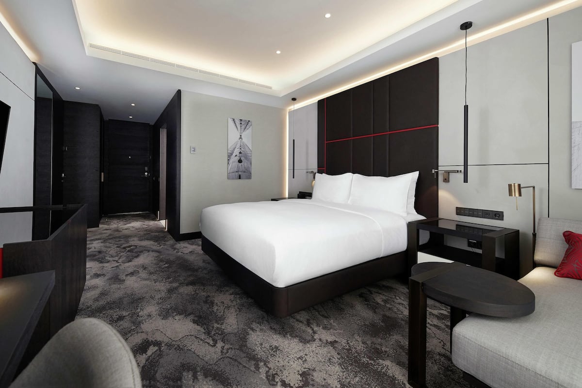 Una habitación de hotel moderna con una cama grande, un escritorio, una silla, un televisor y un cojín rojo decorativo.