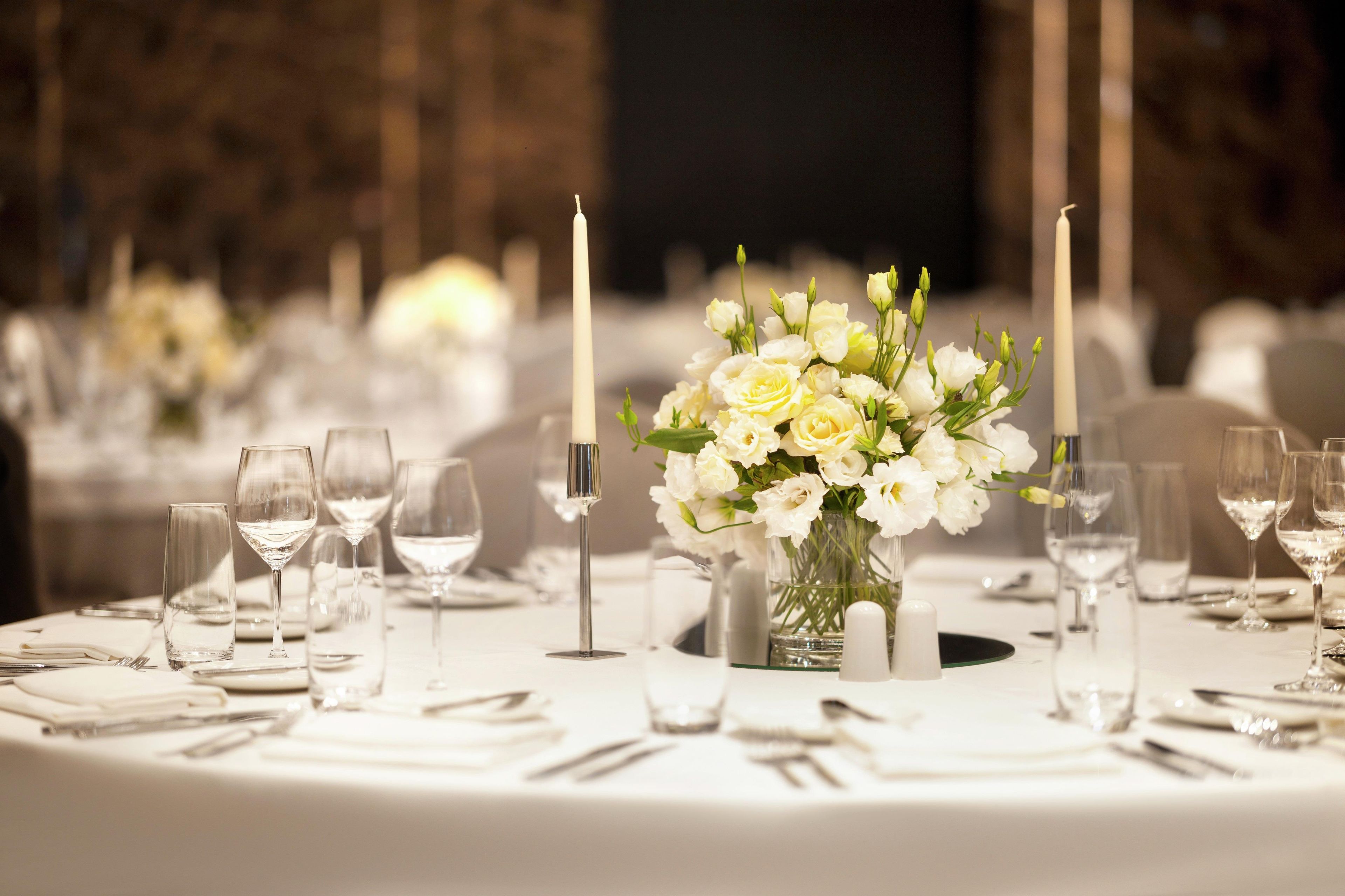 A round table with a white tablecloth, a vase of white flowers, candles, and empty wine glasses.