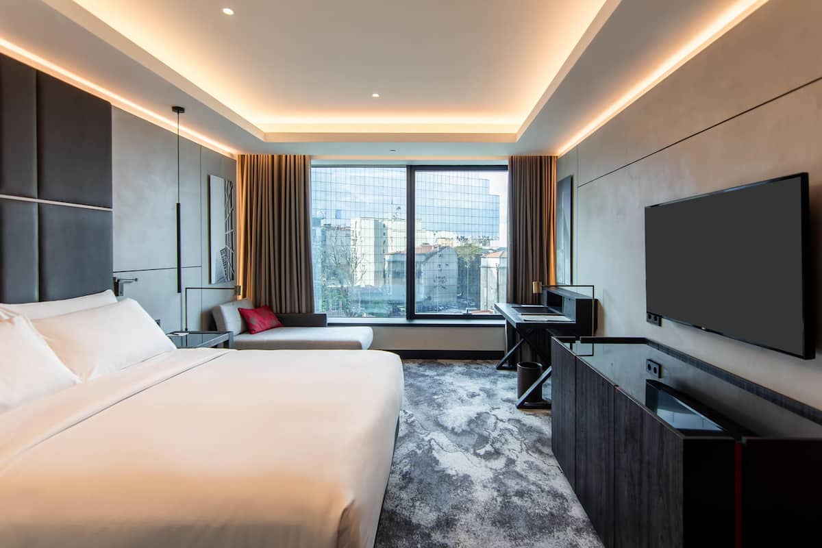 Una habitación de hotel moderna con una cama grande, un escritorio, un televisor de pantalla plana y un ventanal con vistas a la ciudad.