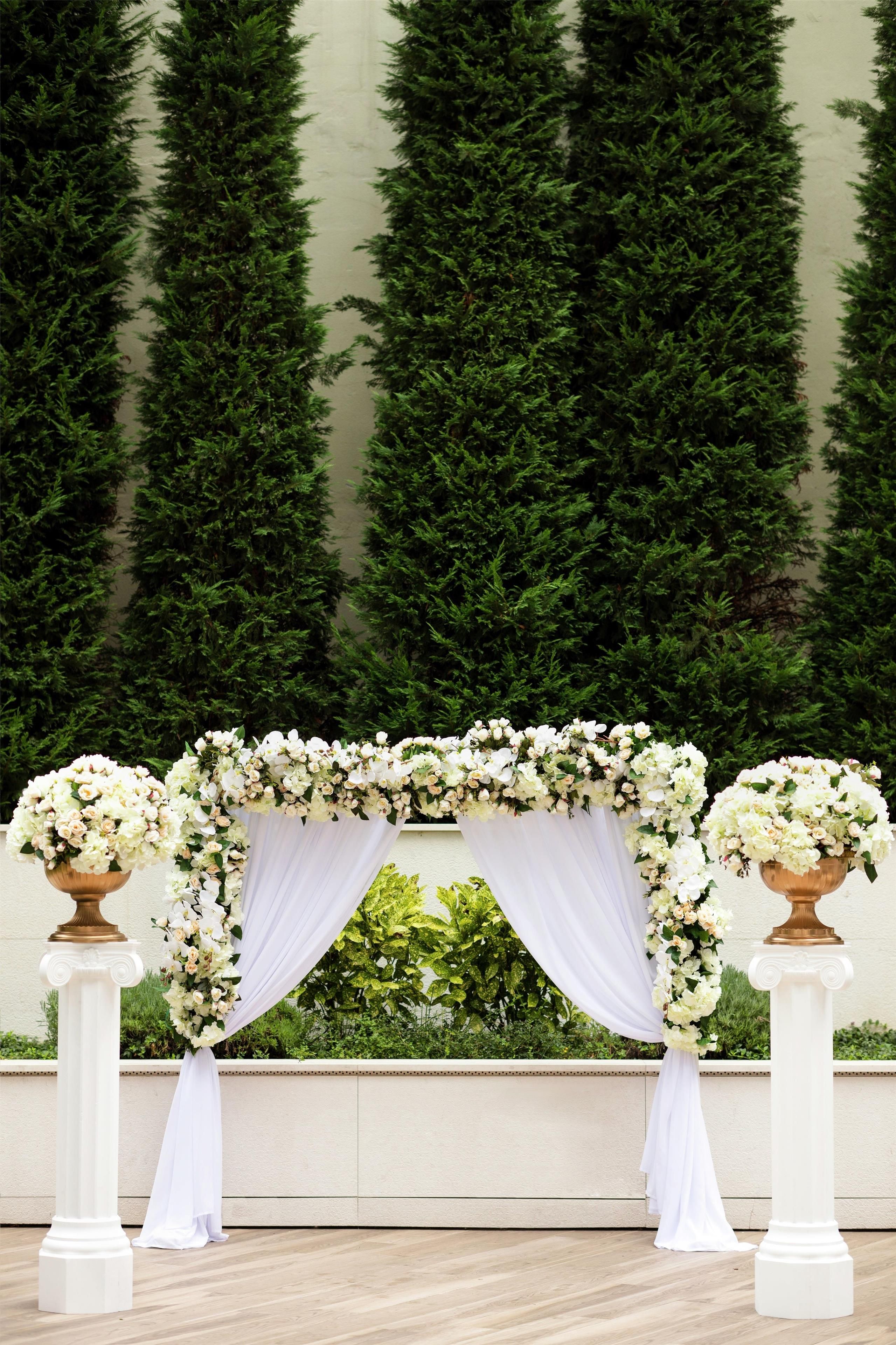 A wedding arch with white flowers and drapes, flanked by white columns, against a backdrop of green foliage.