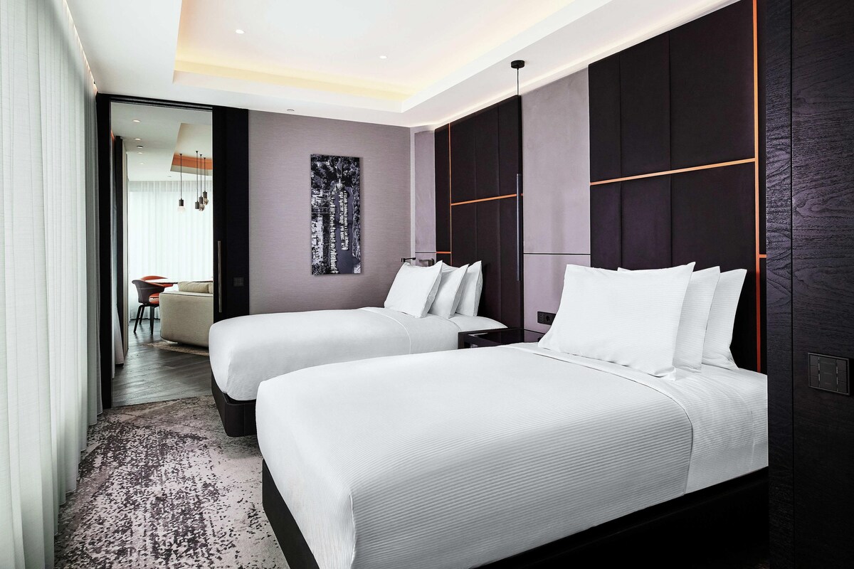 A modern hotel room with two beds, a large abstract artwork on the wall, and a view into another room.