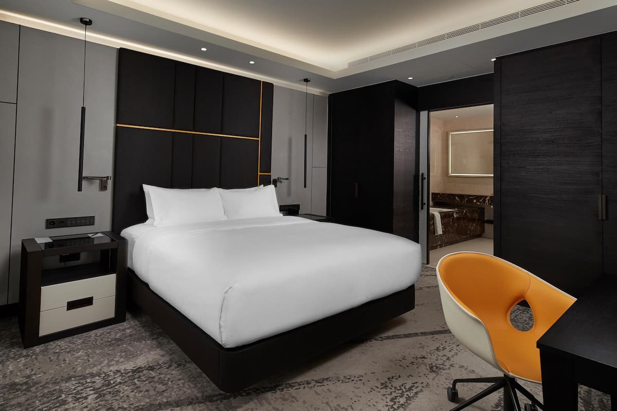 Habitación de hotel moderna con una cama grande, un escritorio, una silla y un baño visible a través de una puerta abierta.