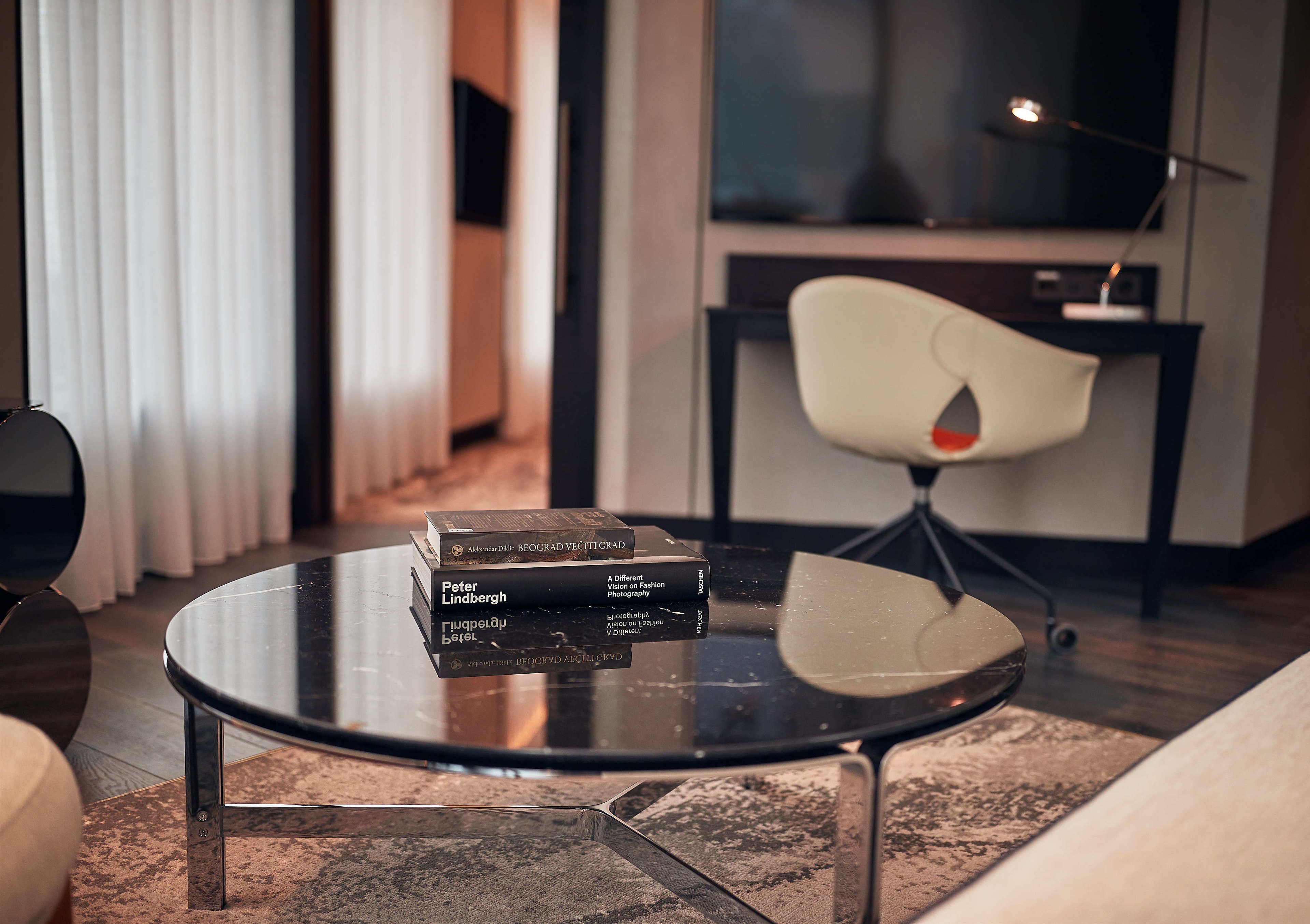 A modern living room with a round glass coffee table, a stack of books, a white chair, and a sofa.