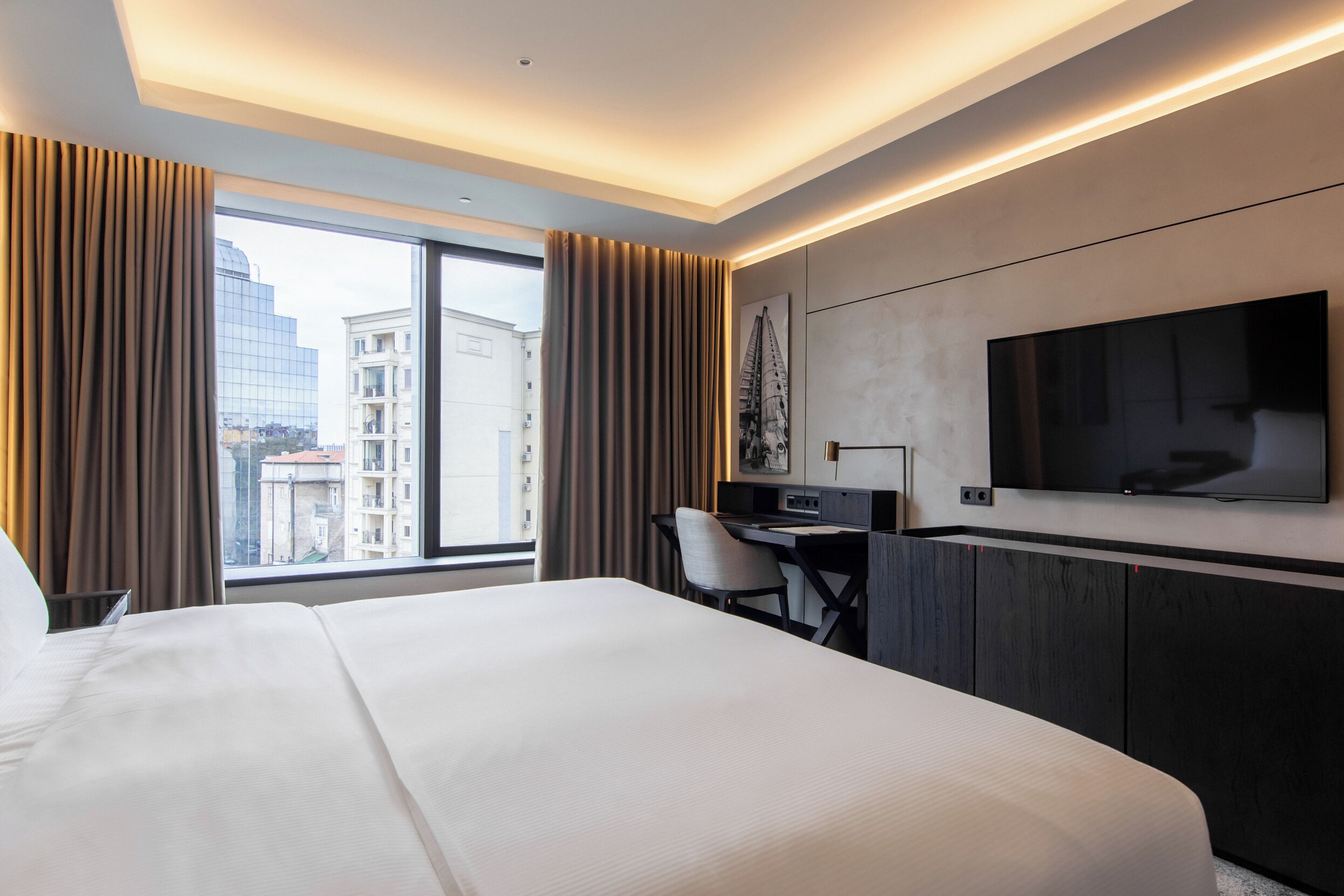Habitación de hotel con una cama grande, un escritorio, una silla, un televisor de pantalla plana y vistas a edificios a través de la ventana.