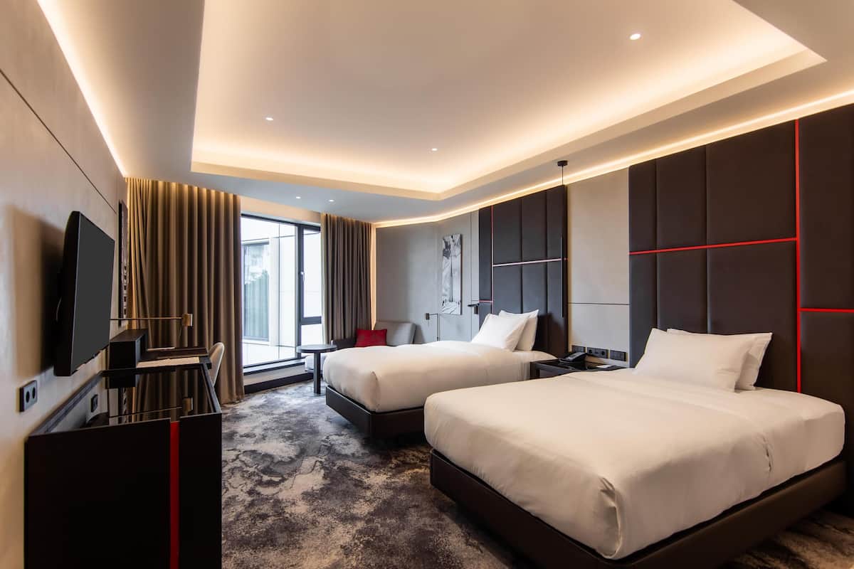Habitación de hotel con dos camas, televisor, escritorio y ventana grande.