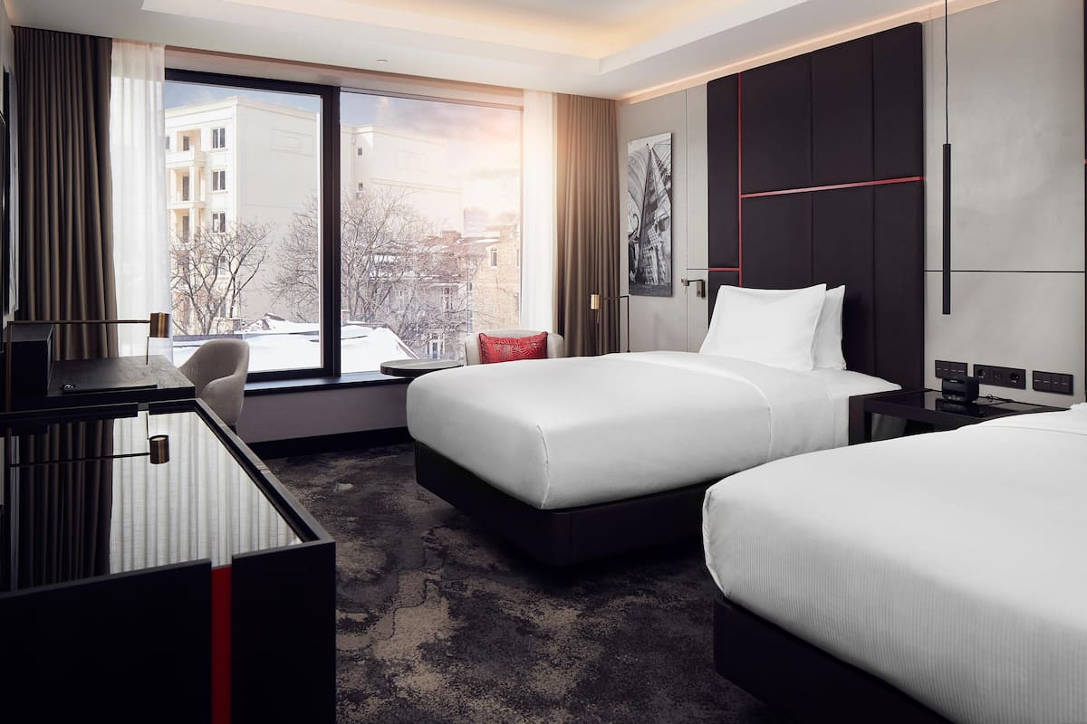 Habitación de hotel con un ventanal grande, un escritorio, dos camas y vistas a edificios.