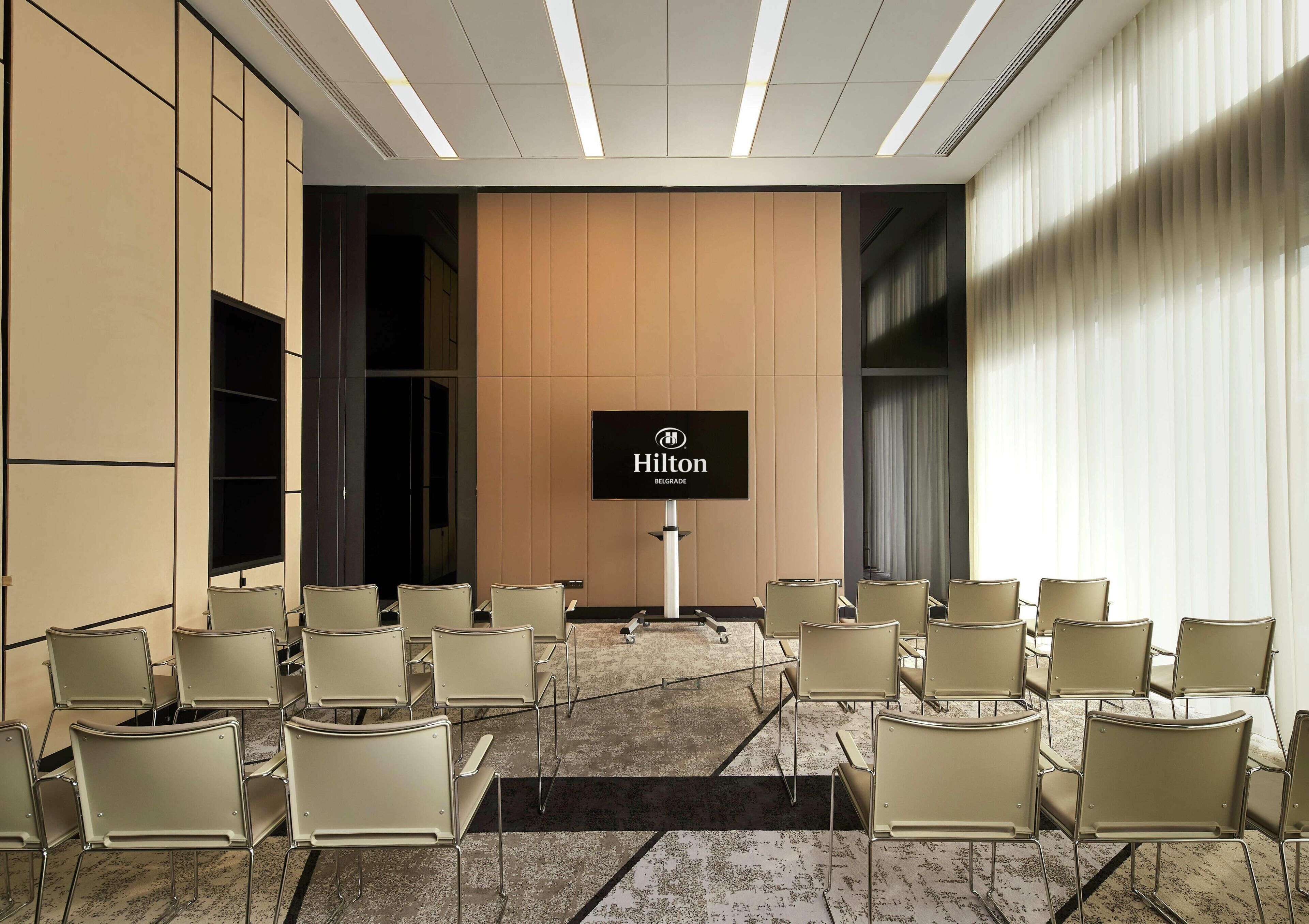 A conference room with a large screen displaying the Hilton logo, beige chairs arranged in rows, and a carpeted floor.