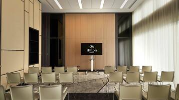 A conference room with a large screen displaying the Hilton logo, beige chairs arranged in rows, and a carpeted floor.