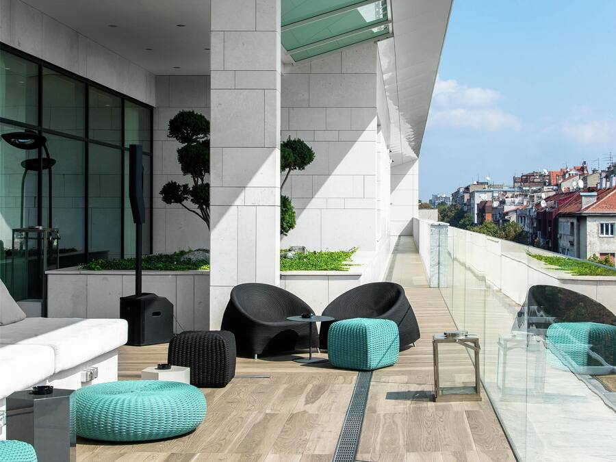 A modern outdoor lounge area with seating, a glass railing, and a view of the city.
