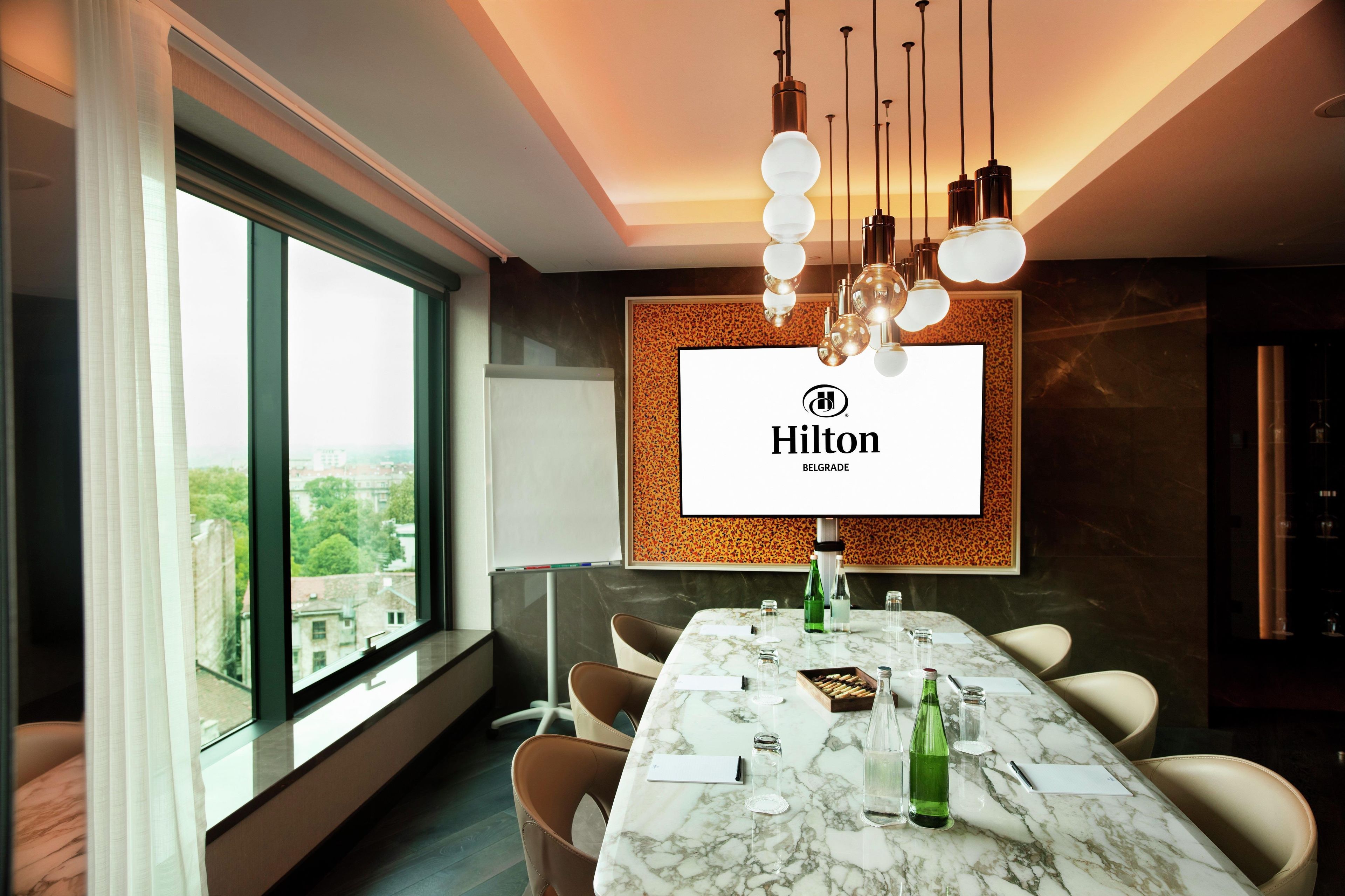 A modern hotel conference room with a large table, comfortable chairs, a large window, and a branded screen.