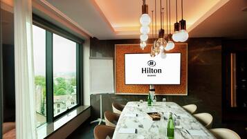 A modern hotel conference room with a large table, comfortable chairs, a large window, and a branded screen.