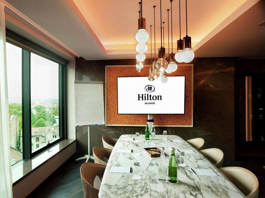 A modern hotel conference room with a large table, comfortable chairs, a large window, and a branded screen.