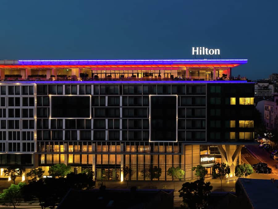 Un hôtel à plusieurs étages, reconnaissable à son grand panneau « Hilton », doté d’un bar sur le toit et arborant un design architectural moderne.