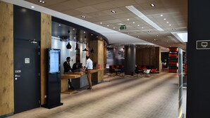 A modern reception area with a wooden reception desk, a digital display, and a seating area with red and black chairs. - ibis Sfax (Sfax Ouest)