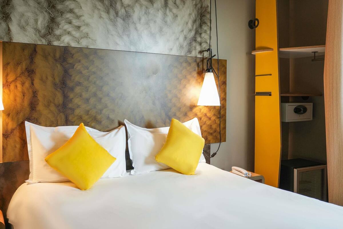 A hotel room with a large bed, two pillows, a headboard with a textured design, a wall-mounted lamp, and a telephone.