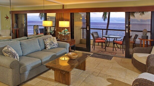 A living room with a sofa, coffee table, and chairs. There is a view of the ocean through large glass doors. - Poipu Shores 303A - Oceanfront Luxury w/ AC & Pool (Koloa)