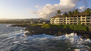 A beachfront hotel with multiple balconies, a rocky shoreline, and palm trees. - Poipu Shores 303A - Oceanfront Luxury w/ AC & Pool (Koloa)