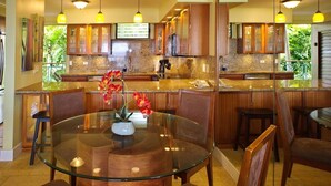A modern kitchen with granite countertops, wooden cabinets, and a glass dining table with chairs. - Poipu Shores 303A - Oceanfront Luxury w/ AC & Pool (Koloa)