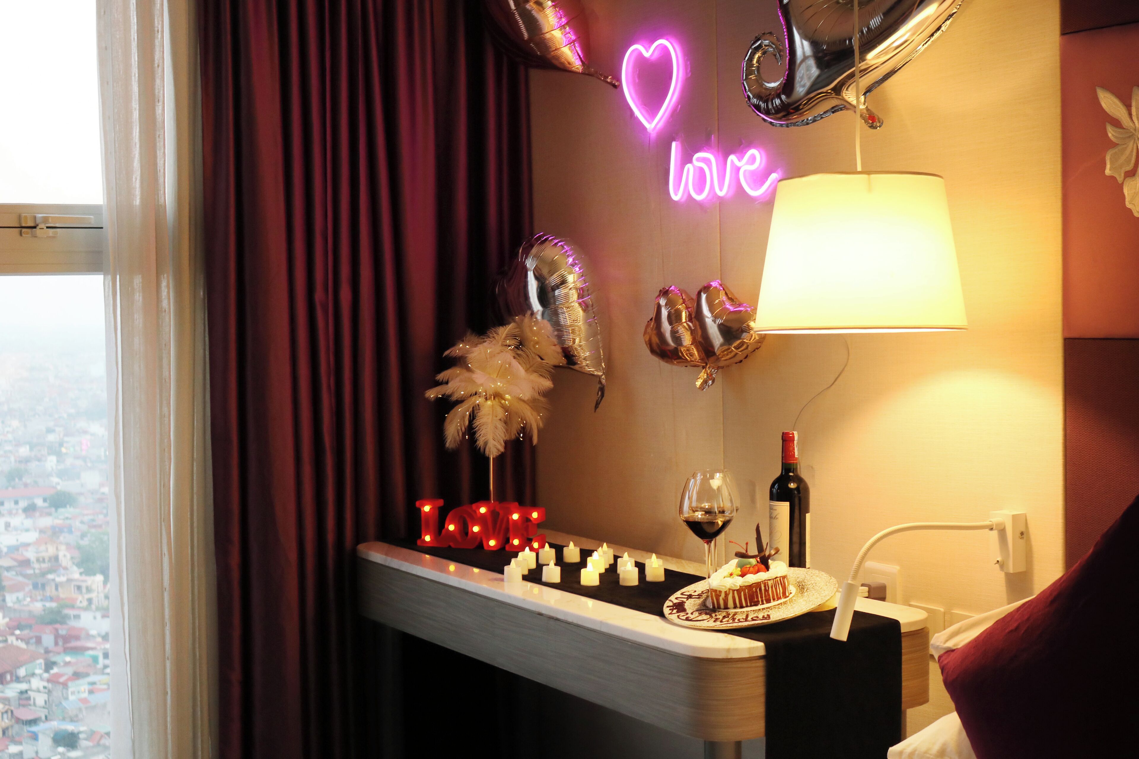 Une chambre avec vue sur la ville, un néon en forme de cœur, des ballons, des bougies et une bouteille de vin posée sur une table.