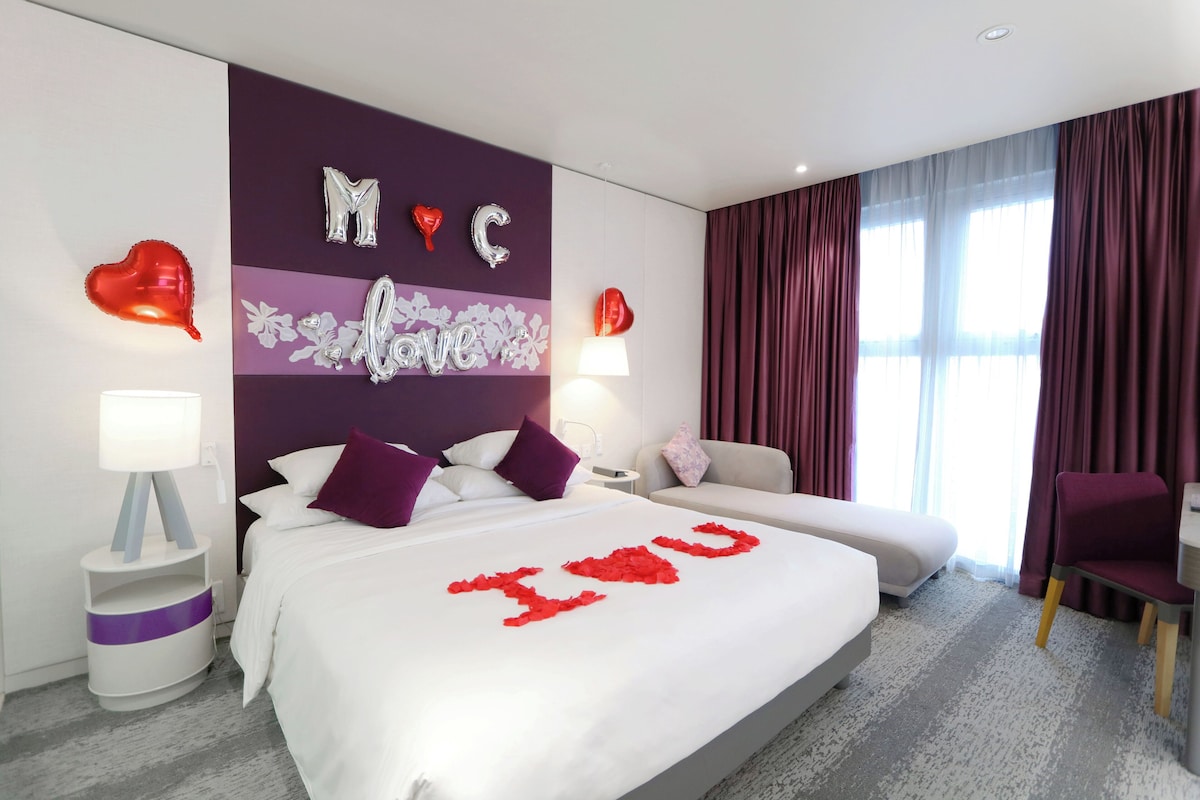 A hotel room with a bed, a sofa, a chair, and a desk. There are decorative pillows and a wall with a heart-shaped decoration.