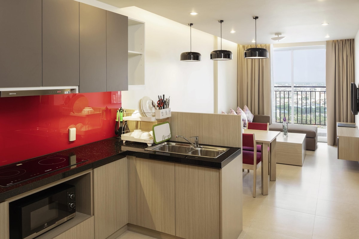 Een moderne keuken met een rode achterwand, houten kastjes en een ingebouwde magnetron. Er is een eethoek met een tafel en stoelen, en een groot raam met uitzicht op de stad.