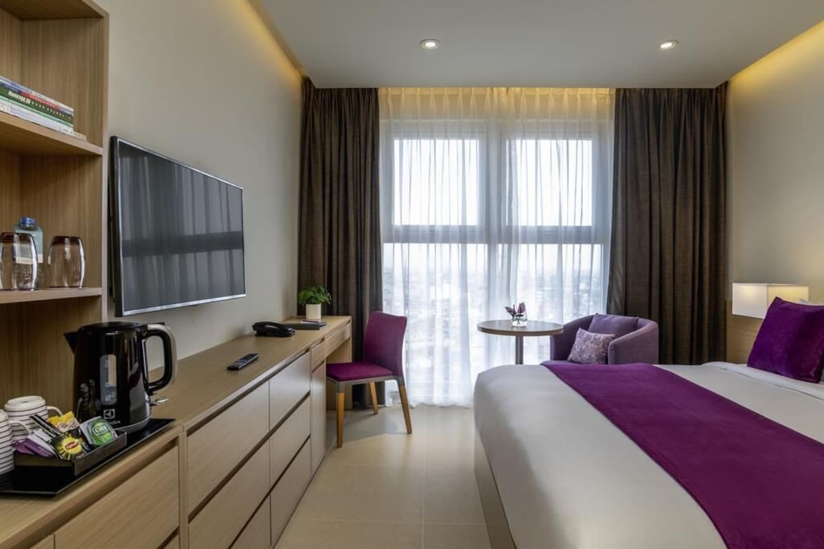 A hotel room with a bed, a desk with a TV, a coffee maker, a chair, a small table, and a window with curtains.