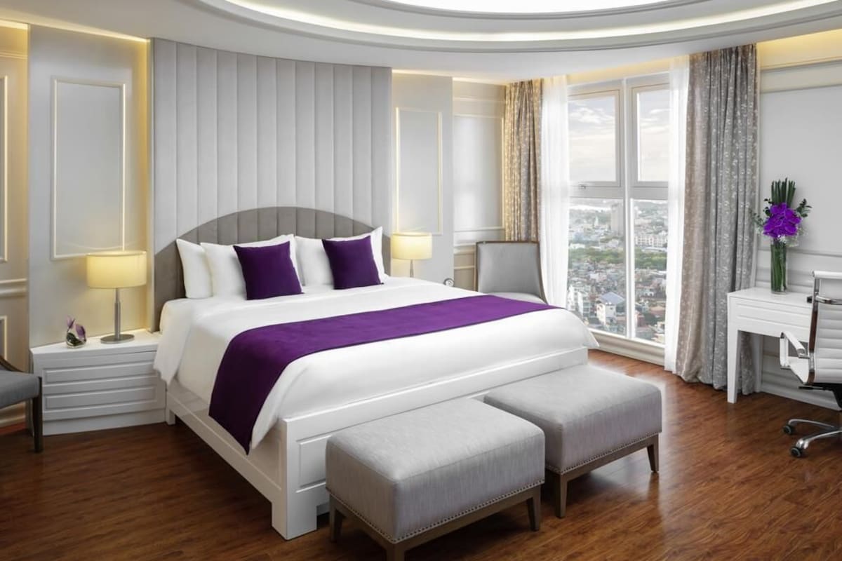 A modern hotel room with a large bed, a bench, a desk, and a view of the city.