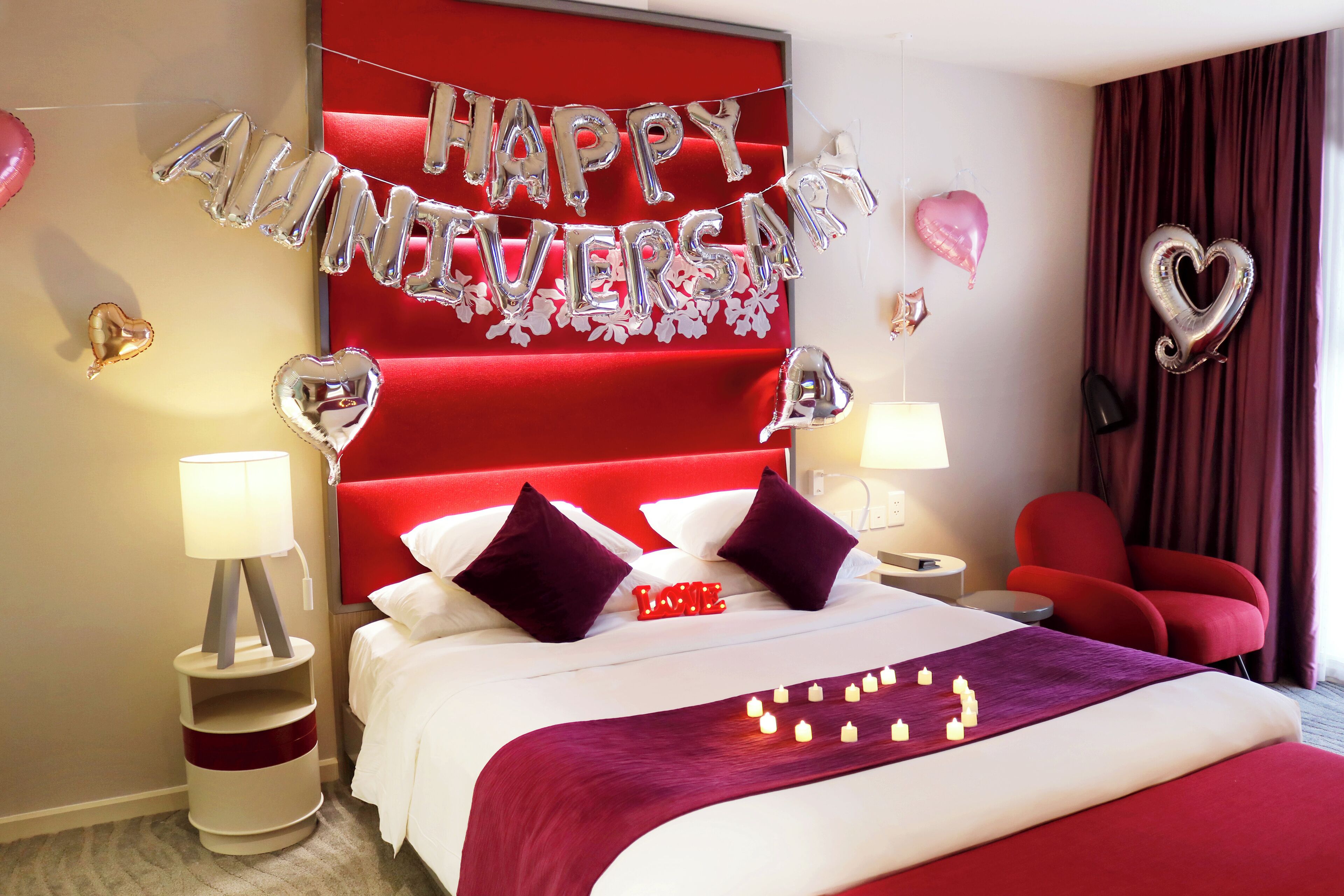 A hotel room with a red headboard, a bed with white bedding, two red chairs, and a 'Happy Anniversary' balloon decoration.