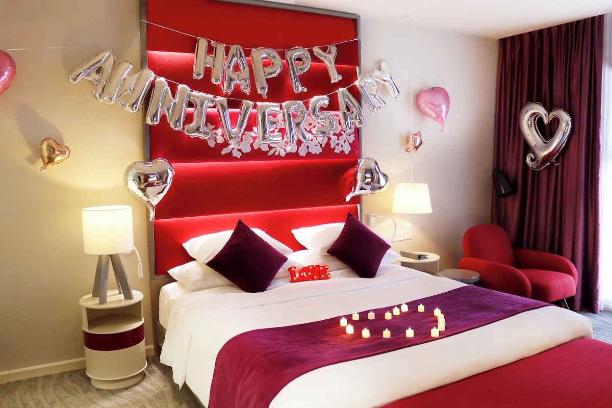 A hotel room with a red headboard, a bed with white bedding, two red chairs, and a 'Happy Anniversary' balloon decoration.