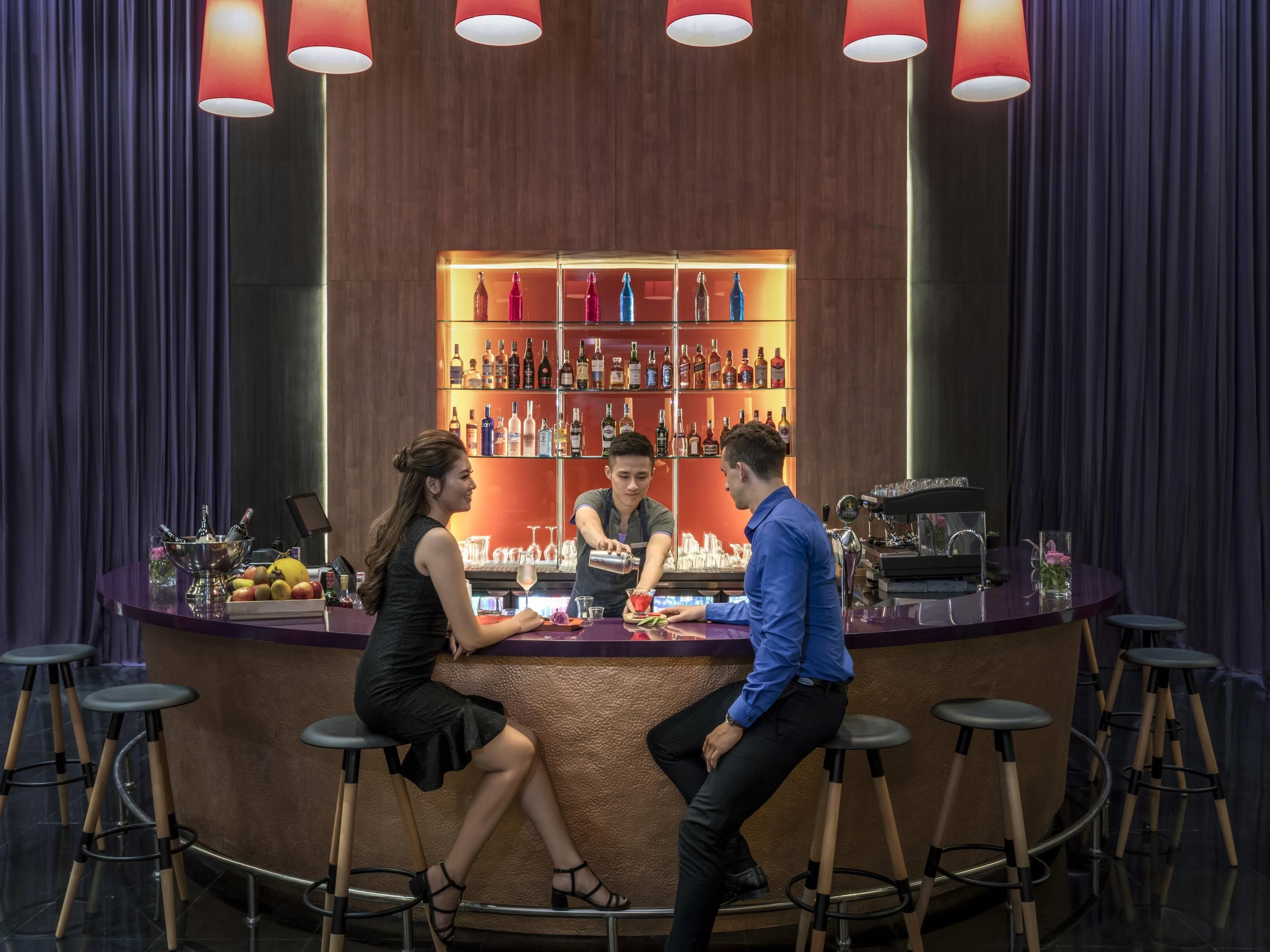 En bardisk med en bartender som blandar drinkar, en sittande kvinna och en man i en blå skjorta.