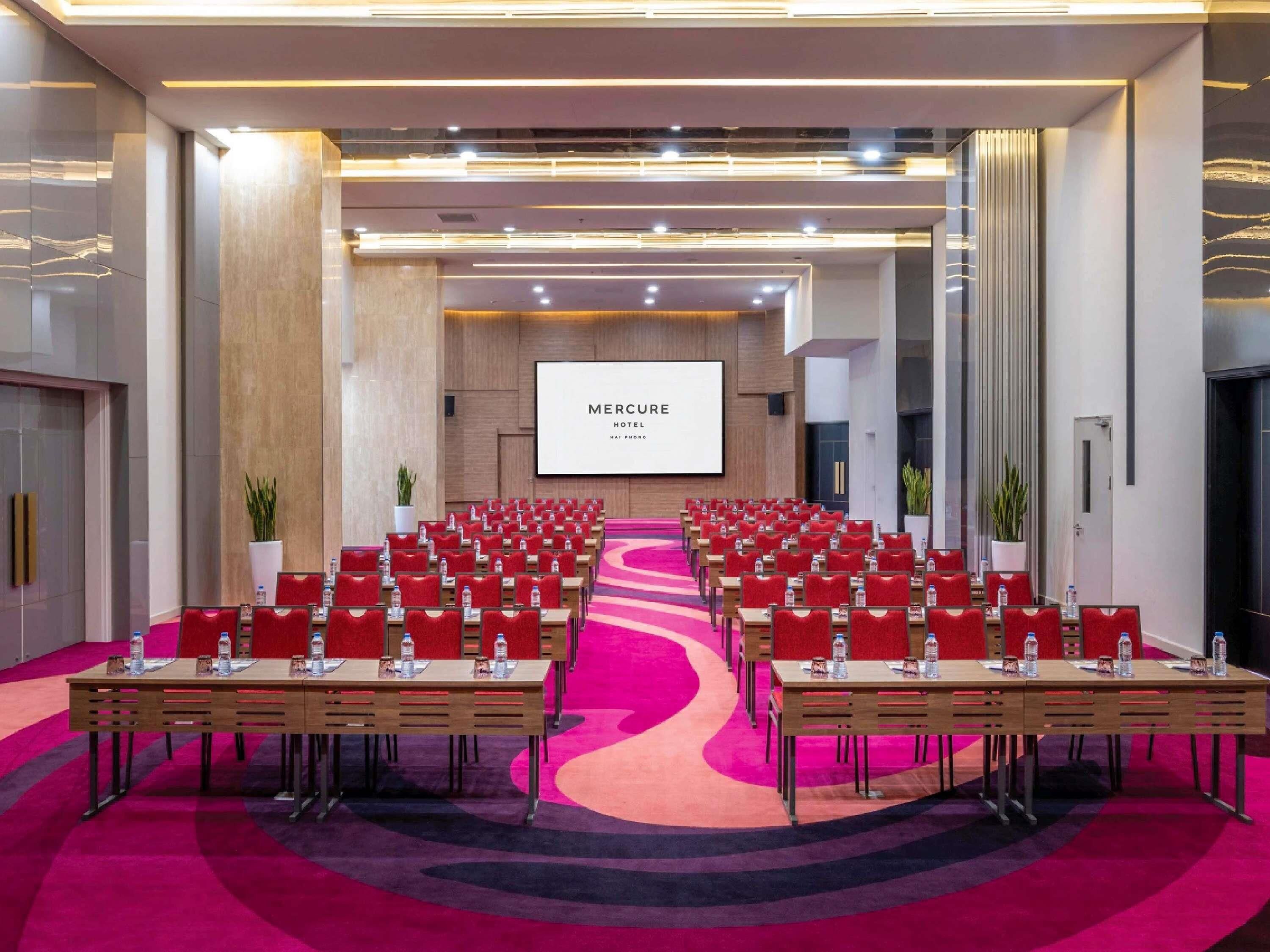 A conference room with rows of tables and chairs, a large screen displaying 'MERCRE HOTEL,' and a carpeted floor.
