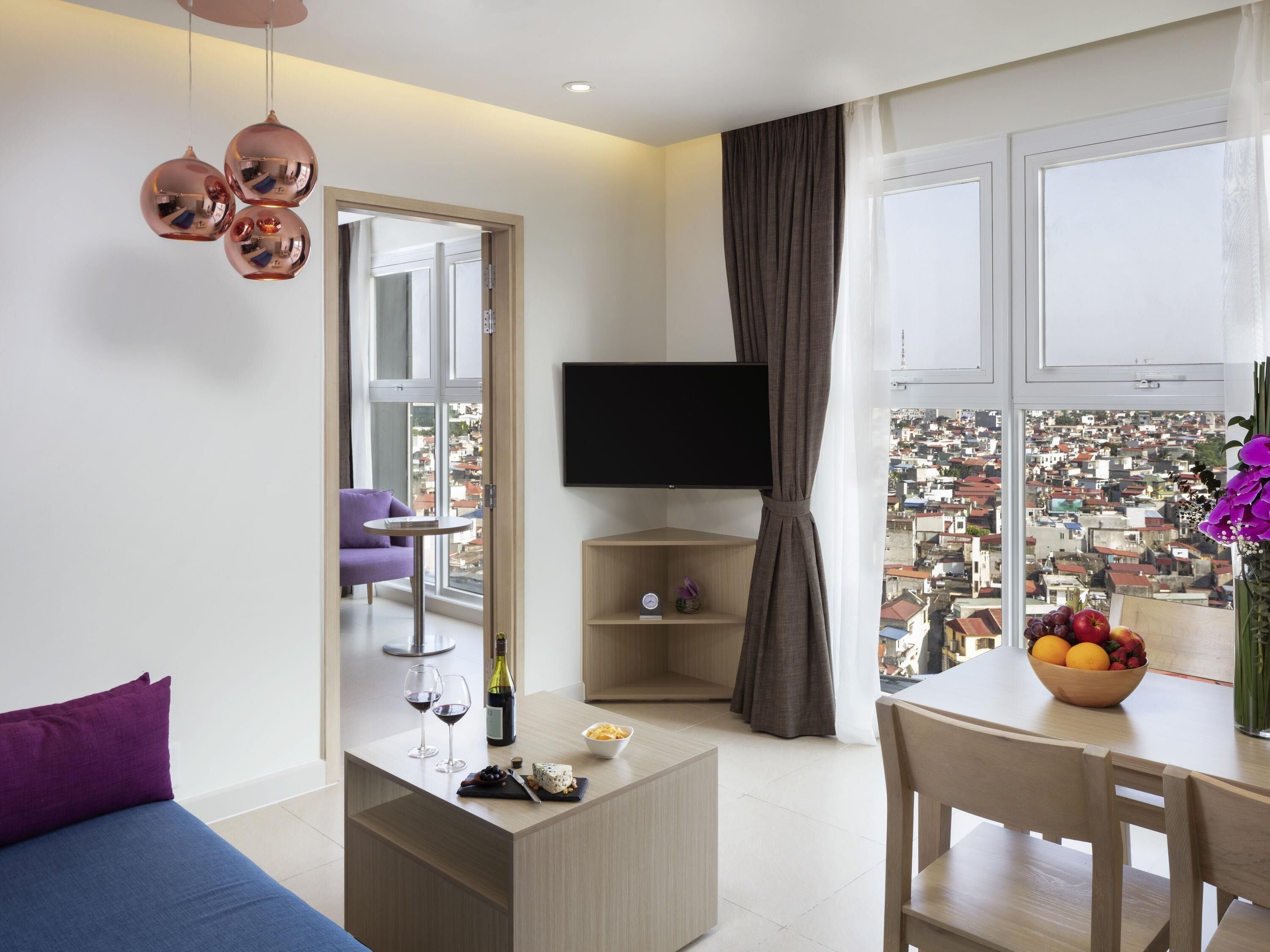 Ein modernes Wohnzimmer mit einer Couch, einem Couchtisch mit Weingläsern und Käse, einem Fernseher, einem Essbereich mit Obstschale und Blick auf die Stadt durch große Fenster.