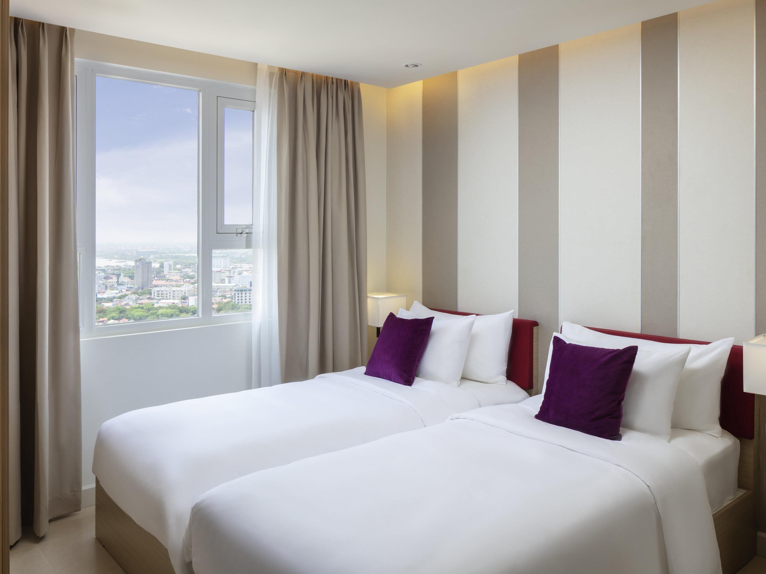 Ein Hotelzimmer mit zwei Betten, einem großen Fenster mit Vorhängen und Blick auf die Stadt.