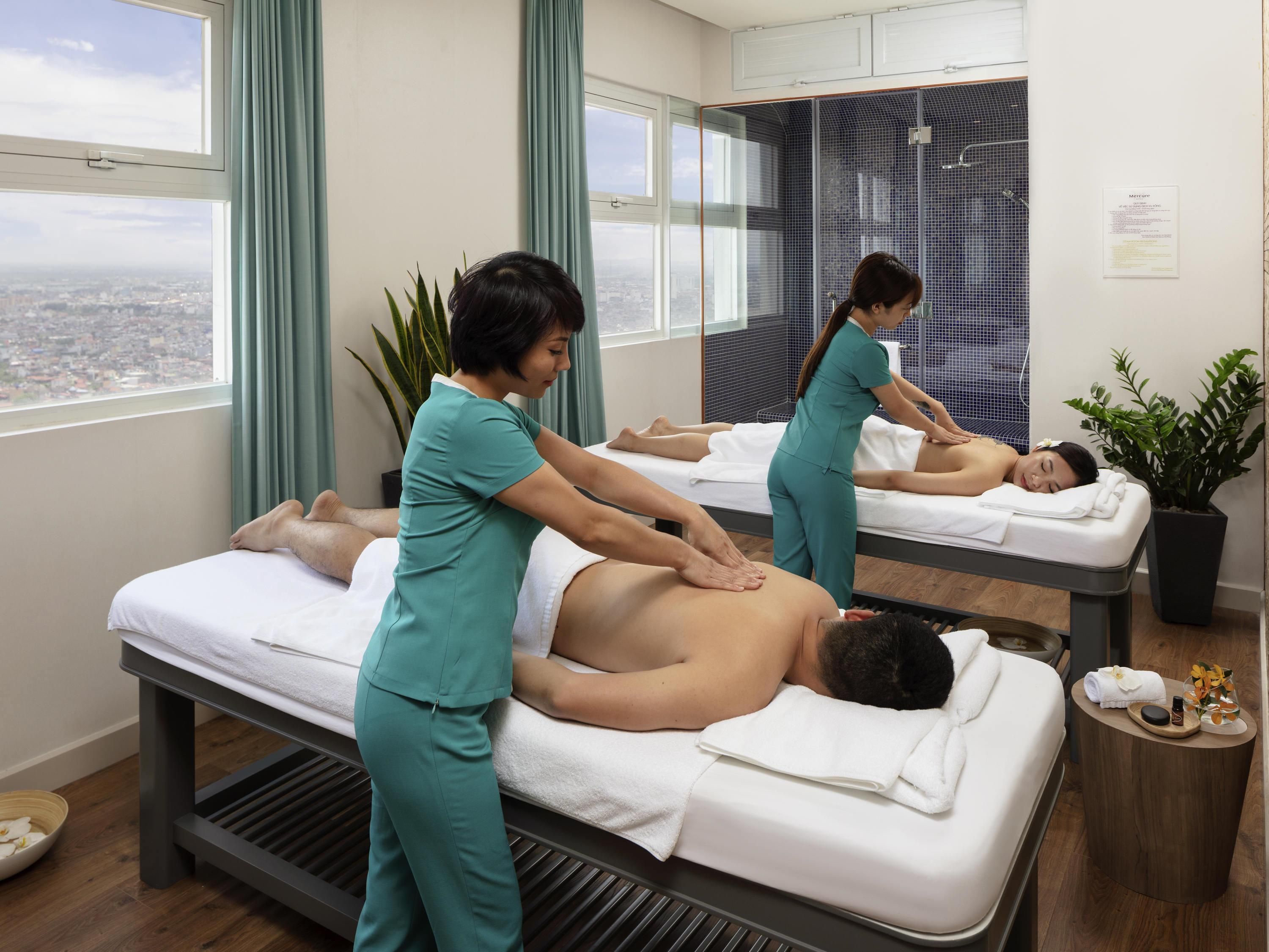 Un cuarto de terapia de masajes con dos camillas, un ventanal con vista a la ciudad y una planta en maceta.