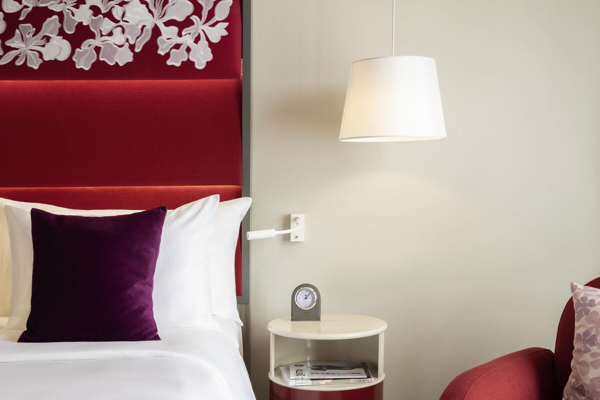 A hotel room with a red upholstered headboard, white bedding, a purple pillow, a white lamp, and a bedside table with a clock and books.