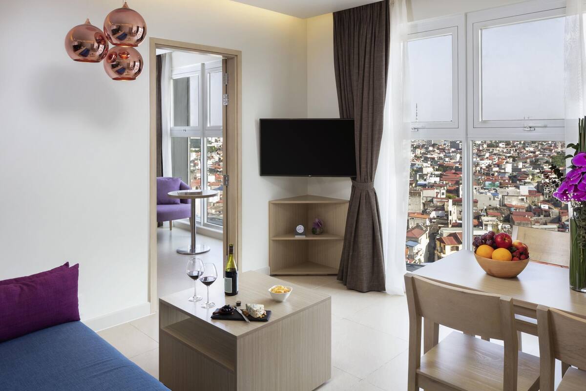 Een moderne woonkamer met een bank, een bijzettafeltje met wijnglazen en kaas, een televisie en een stadsgezicht door grote ramen.