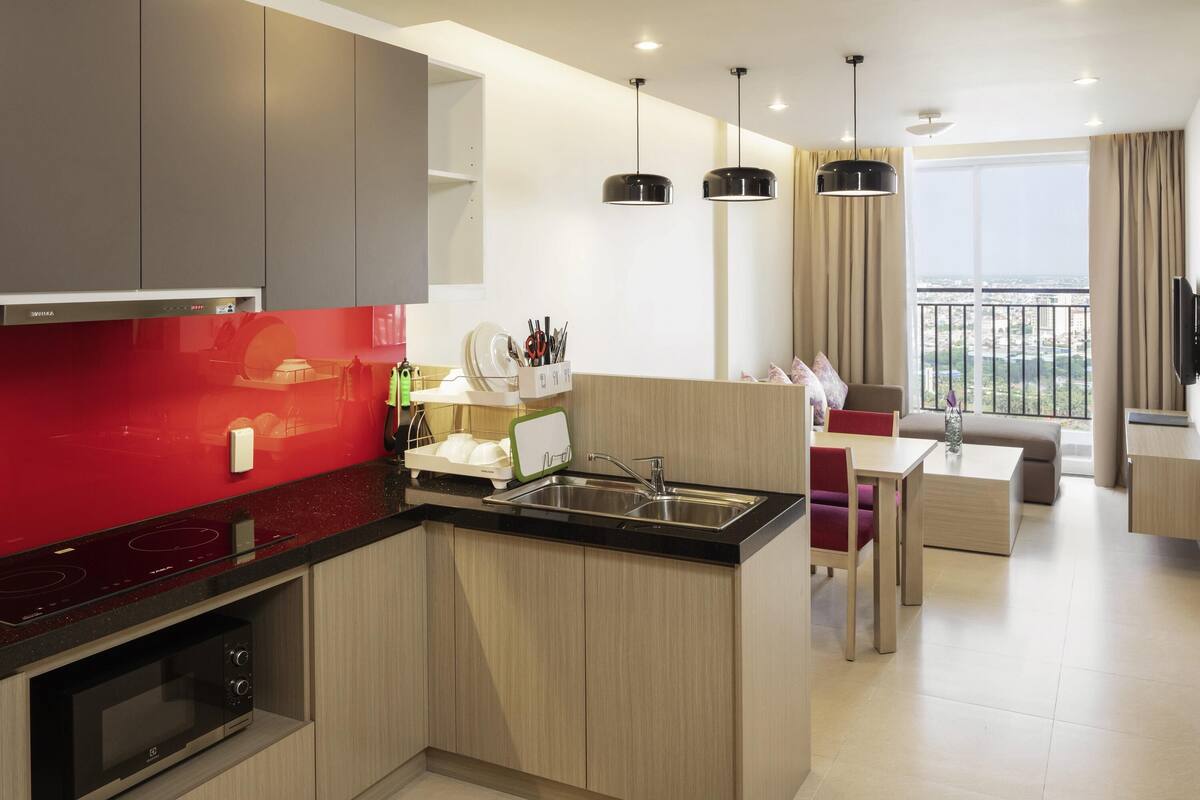 Een moderne keuken met een rode achterwand, houten kastjes en een ingebouwde magnetron. Er is een eethoek met een tafel en stoelen, en een groot raam met uitzicht op de stad.
